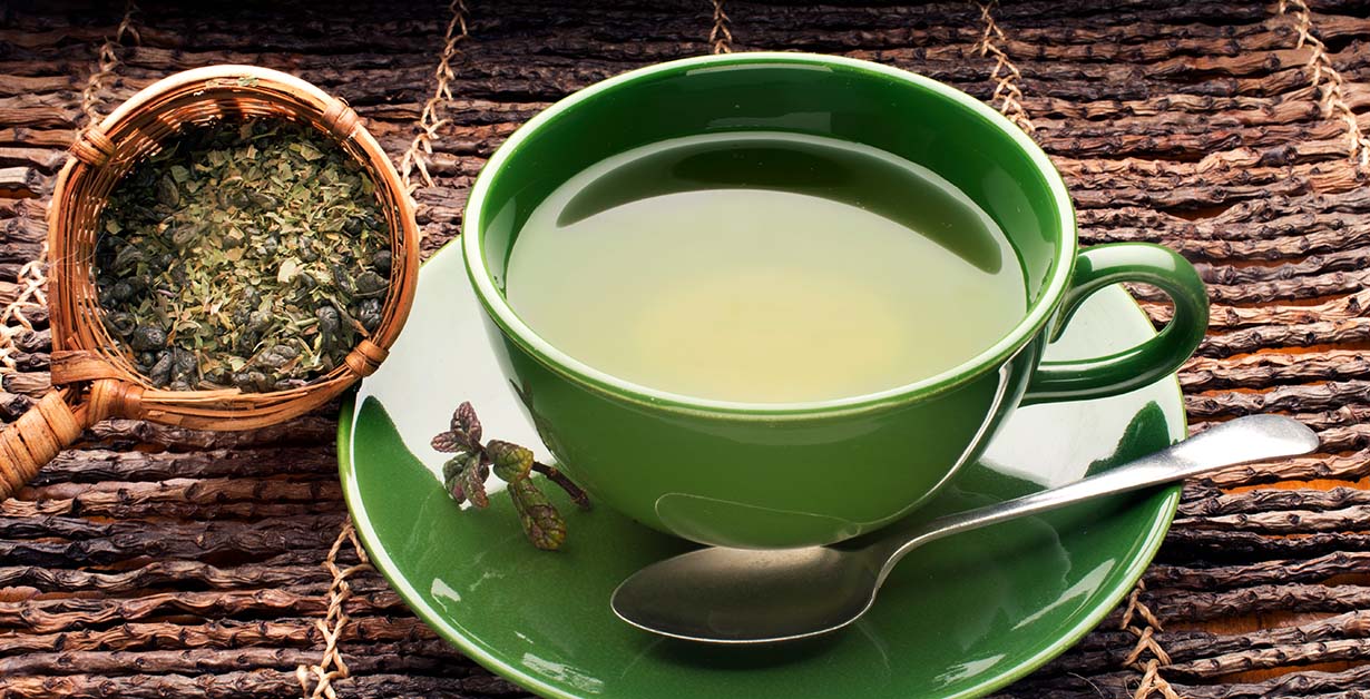 A spoon of green tea leaves next to a cup of green tea.